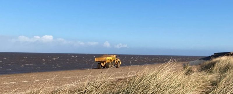 Yellow dumper truck moving along a sandy beach with a bright blue sky behind. Photo by Lizzie Grayshon