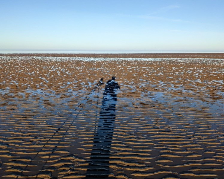 A shadow of someone standing next to a scope on a tripod resighting birds. The shadow is falling across a beach with ripples filled with water and reflecting a clear blue sky. Photo by Rob Robinson.
