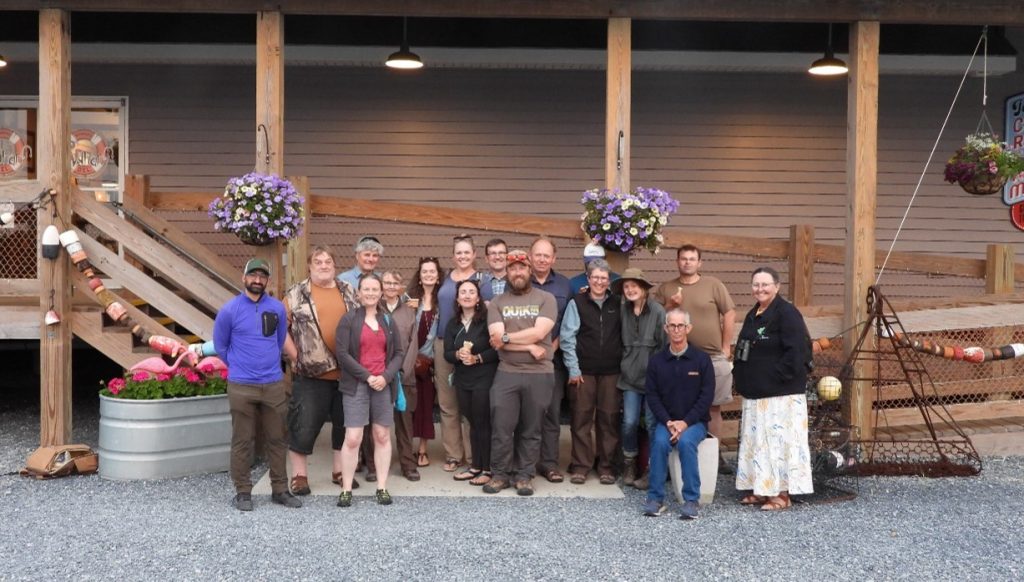 Group photo of the team members outside a restaurant. There are hanging baskets, tubs with flowers and fishing equipment decorating the outside of the wooden building. Photo by Cathy Ryden.