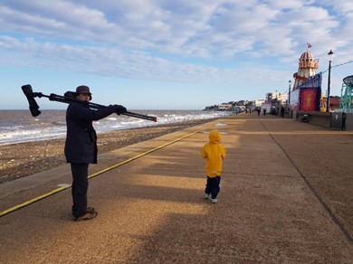 Photo of a man and a child walking along a promenade. The man is carrying a spotting scope over his shoulder. The child is in a bright yellow jacket, with the hood up.